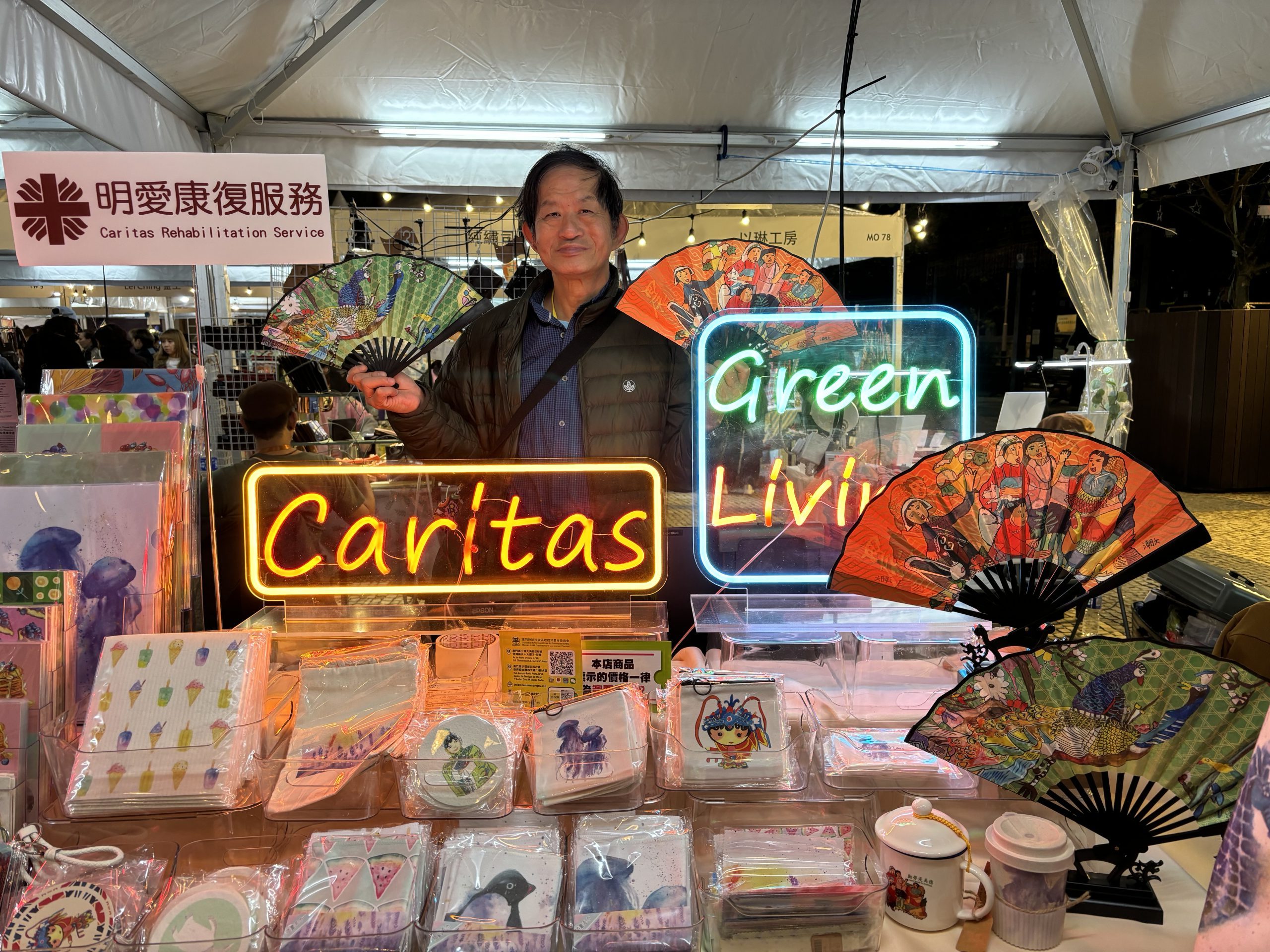 兩位老闆Mini和老闆潮文當值於澳門塔石藝墟攤位內，周圍擺放著多款手工藝產品，展現出熱鬧的市集氛圍。