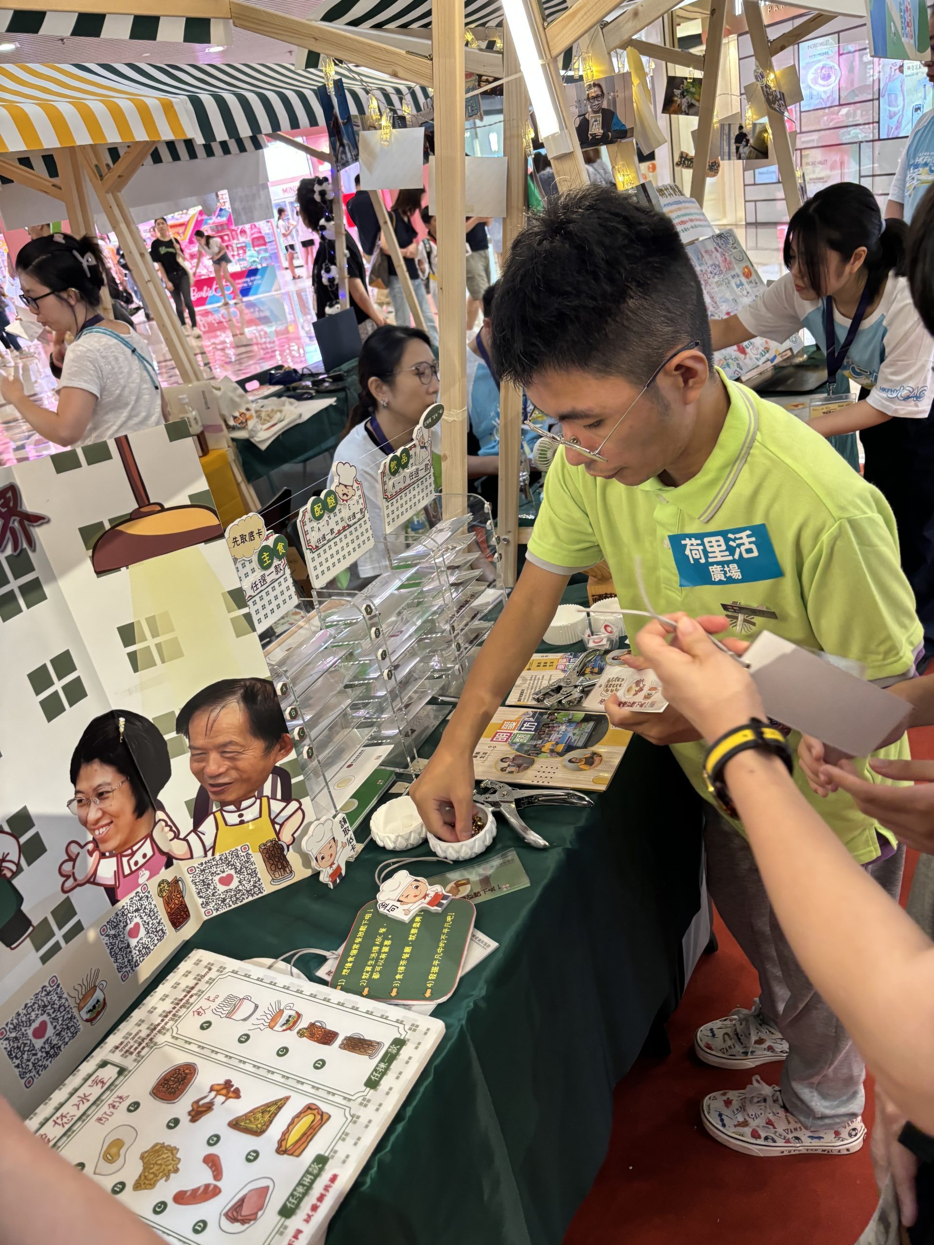 老闆健仔站在桌子旁，向社區人士介紹如何製作茶餐廳書籤。
