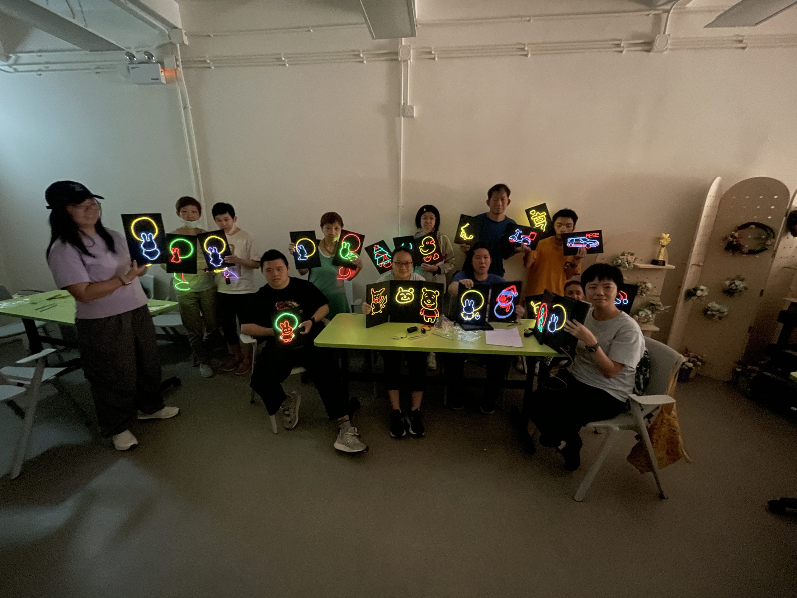 組員們舉著發光的冷光線（霓虹燈）牌，燈光閃爍，氣氛熱烈，展示著大家的作品成果。