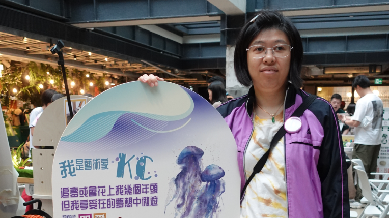 A woman is holding a sign featuring her own artwork, which displays a jellyfish painting, reflecting her satisfaction and happiness.