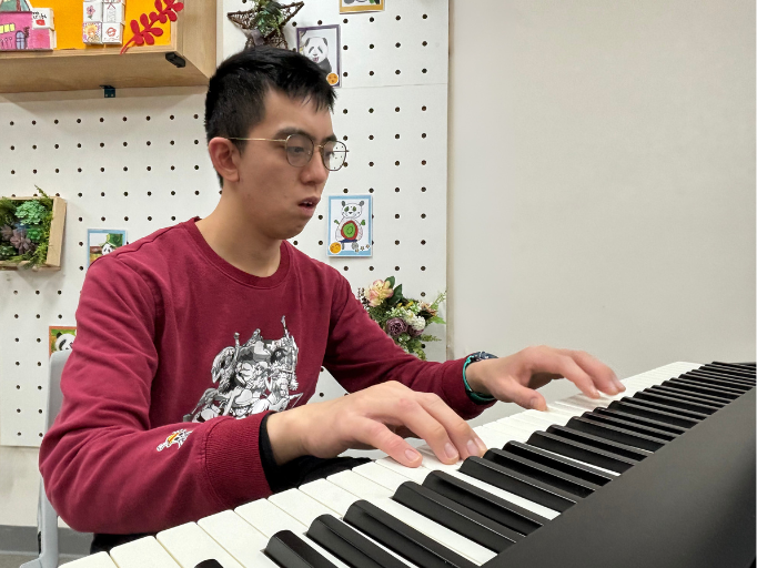 Artist Bon, wearing a red long-sleeve shirt, plays the piano with a focused expression, immersed in his own musical world.