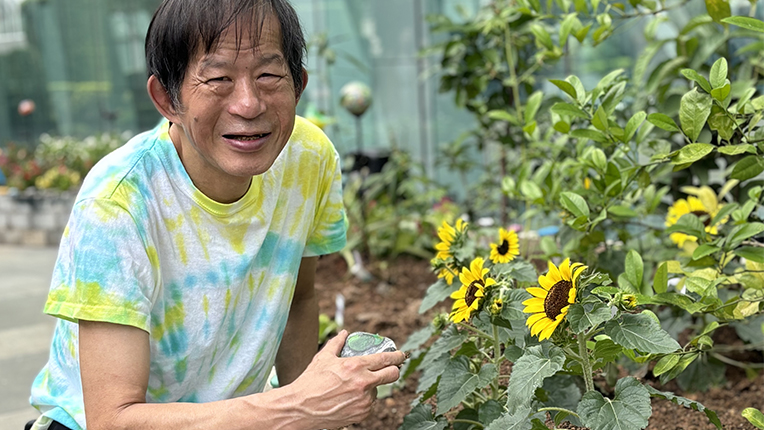 Boss Man is wearing a tie-dye shirt and holding a sunflower, displaying a sunny smile.